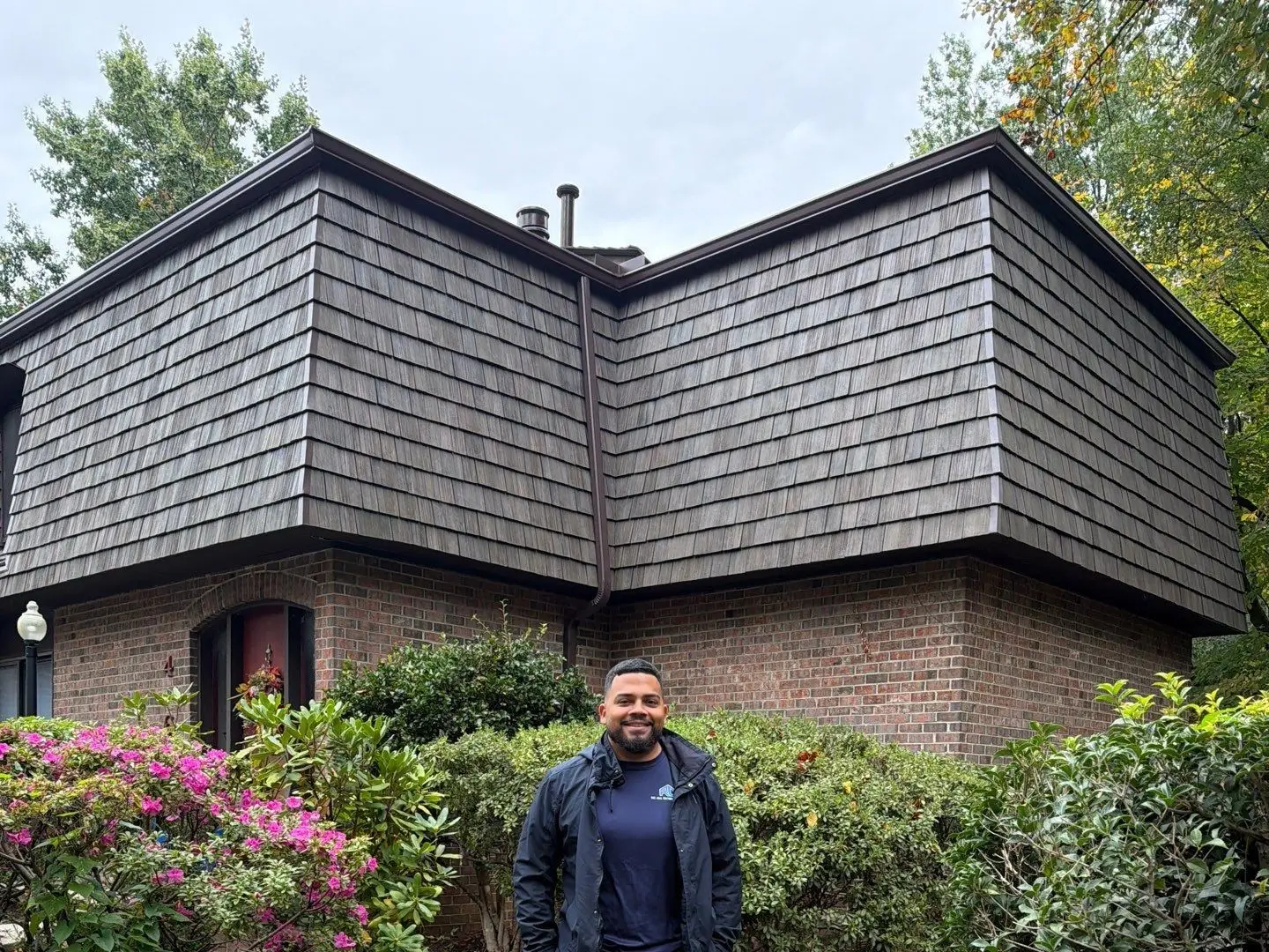 An RC All Exteriors professional in front of a brick home with its new Brava synthetic shake mansard roof and the RC All Exteriors yard sign